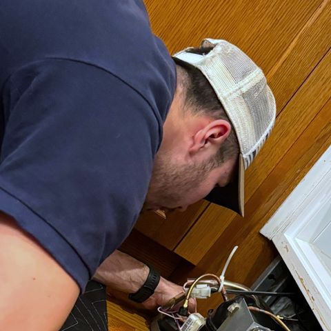 A technician in a cap leans over the opened base of an under-counter refrigerator, reaching into its wiring