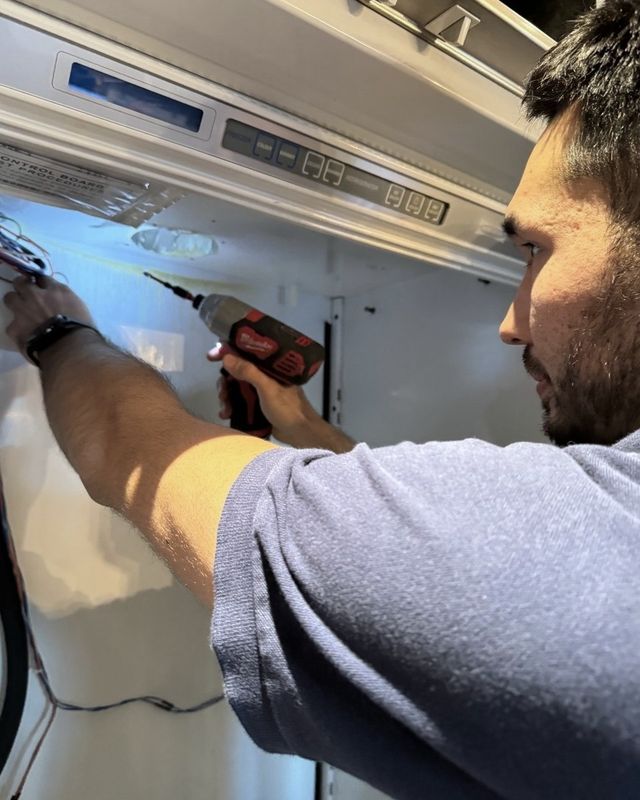 A technician with a cordless driver working at the control panel along the top of a built-in refrigerator