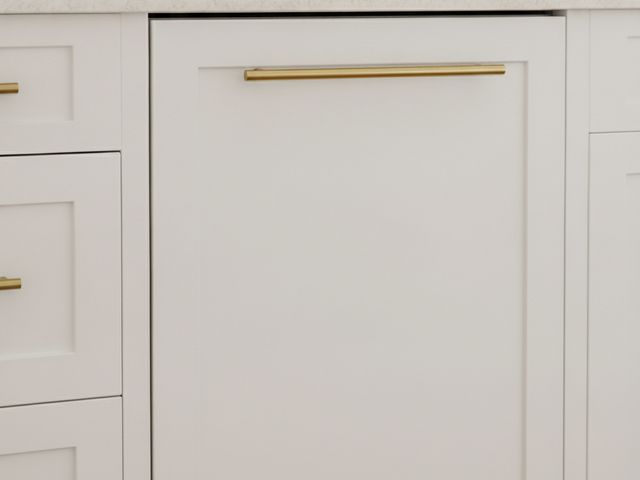 A dishwasher behind a white shaker cabinet panel with a brass pull, matching the drawers beside it