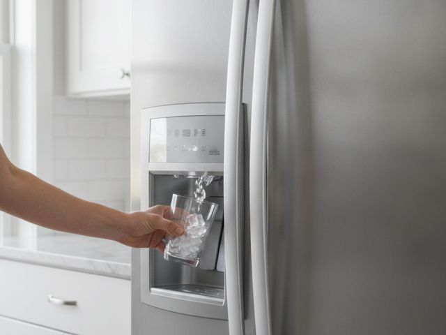 A hand holding a glass to the water and ice dispenser in a stainless refrigerator door
