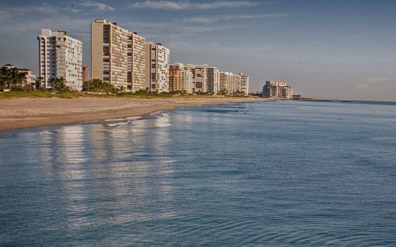 A row of condo towers behind a wide sandy beach, seen across calm morning water