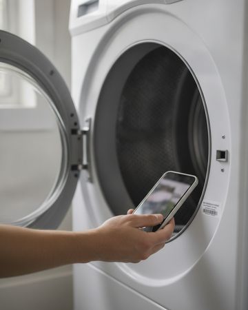 A hand holding a phone to the rim of an open dryer door opening to photograph its label