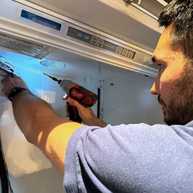 A technician with a cordless driver works at the control panel inside the top of a refrigerator