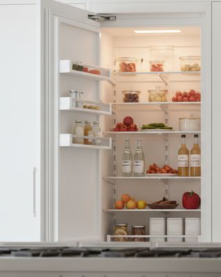 A white panel-ready built-in column refrigerator standing open in a bright kitchen, its shelves stocked