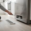 A hand reaching a long, soft brush under a refrigerator, its base grille lying on the floor beside it