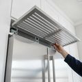 A hand lifting the top grille of a stainless built-in refrigerator, the condenser behind it