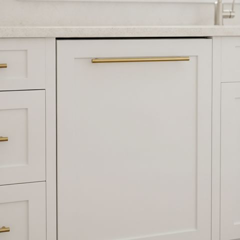 A dishwasher behind a white shaker cabinet panel with a brass pull, matching the drawers beside it