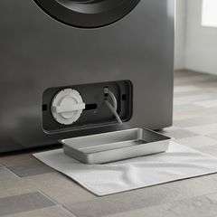 The small access panel at the bottom of a front-load washer open on the drain pump filter cap, a tray and a towel on the floor in front