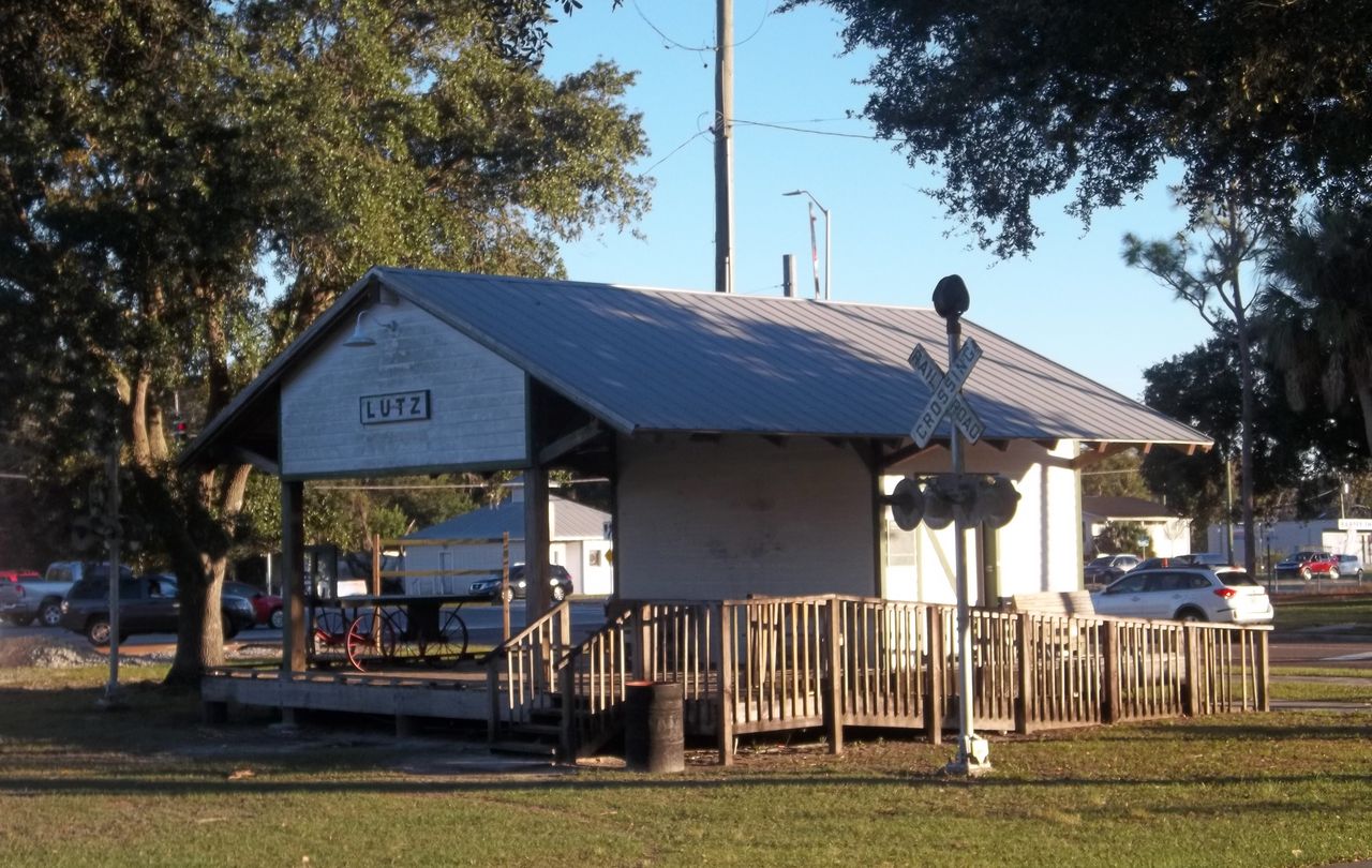 The rebuilt Lutz train depot, its LUTZ sign under a tin roof, beside a railroad crossing signal and live oaks