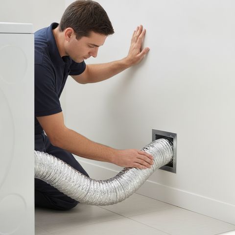 A technician in a navy polo kneeling at a laundry room wall, one hand on the dryer duct where it enters the wall