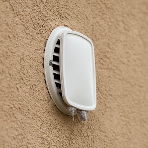 A white dryer vent hood on a stucco wall, a little lint caught under its flap