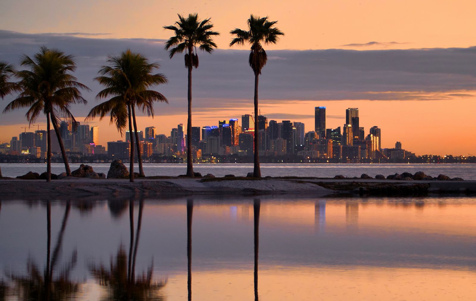The Miami skyline before sunrise under an orange sky, seen across Biscayne Bay past tall palms, all of it reflected in still water