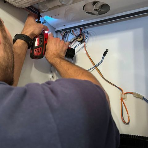 Hands holding a cordless driver beside loose wiring under the control housing of a built-in refrigerator
