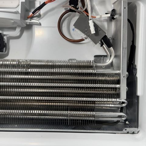 A bare evaporator coil behind a removed panel, its fan motor and defrost wiring above it