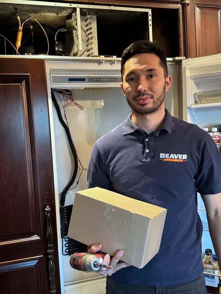 A technician in a Beaver Appliance polo holds a parts box and a cordless driver in front of a built-in refrigerator, its machine compartment open above