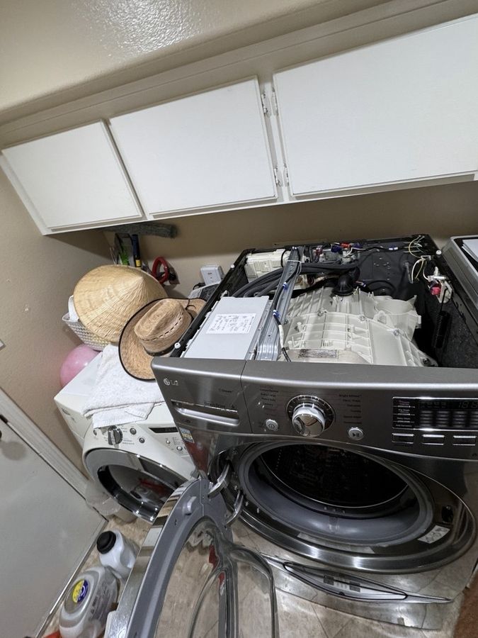 A front-load washer with its top panel off, the tub and wiring open to view, in a laundry room with cabinets above