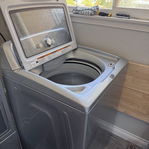 A top-load washer with its lid raised beside a window, screwdrivers on the sill