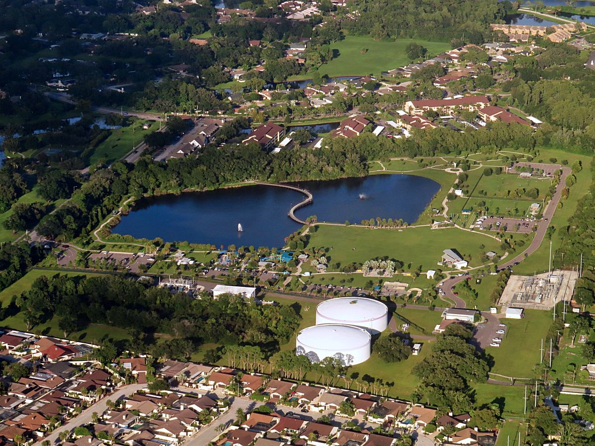Carrollwood from the air: a park pond with a curved footbridge, homes among oaks and two white storage tanks