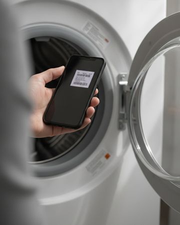 A hand holding a phone at the open door of a front-load washer to photograph its label